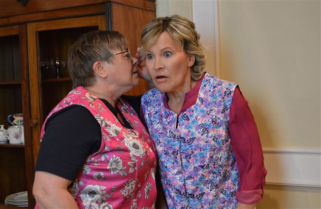 Due donne in grembiuli colorati si stanno fronteggiando e si stanno guardando. Una donna sussurra qualcosa all'orecchio dell'altra, mentre la seconda sembra sorpresa.