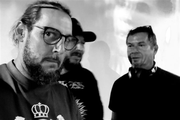 Three men are standing together in an informal setting. They appear relaxed and are conversing.