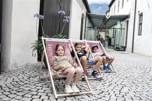 Drei Kinder sitzen entspannt in Liegestühlen auf einem gepflasterten Platz. Im Hintergrund sind Gebäude und Pflanzen sichtbar.