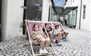 Drei Kinder sitzen entspannt in Liegestühlen auf einem gepflasterten Platz. Im Hintergrund sind Gebäude und Pflanzen sichtbar.