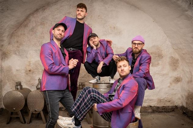 Cinq hommes en vestes colorées posent dans une pièce rustique. L'arrière-plan est constitué d'un mur en pierre et d'objets métalliques.