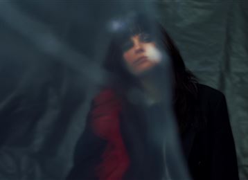 A person with dark hair is standing behind a transparent curtain. The image has a mysterious atmosphere.