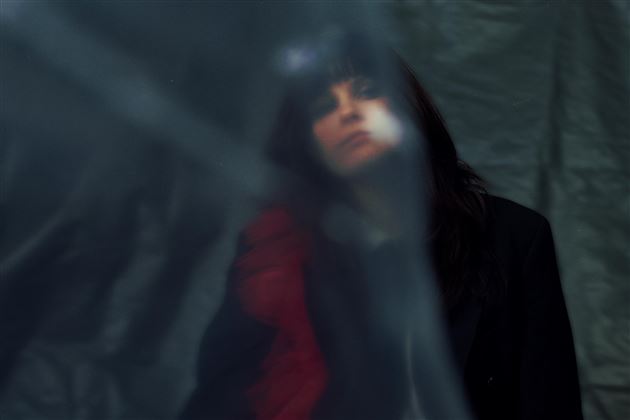 Eine Person mit dunklen Haaren steht hinter einem transparenten Vorhang. Das Bild hat eine mysteriöse Stimmung.