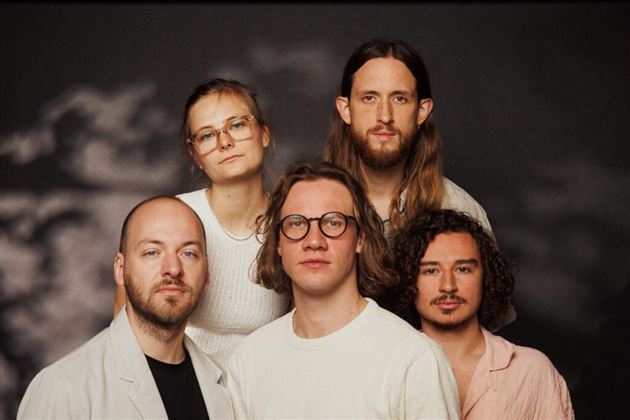 Un gruppo di cinque persone si trova di fronte a uno sfondo scuro. Guardano direttamente verso la fotocamera e indossano diversi vestiti casual.