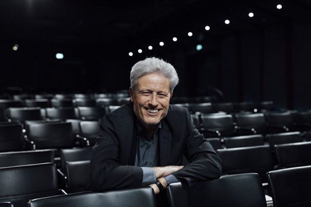 Un homme souriant est assis dans un théâtre vide avec des chaises noires. L'éclairage est doux et l'arrière-plan est sombre.