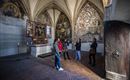 A group of people stands in a historic room with high vaults. The walls are adorned with impressive frescoes and artworks.