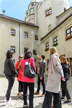Eine Gruppe von Menschen steht in einem Innenhof und hört einem Führer zu. Alte Gebäude mit Fenstern und Pflanzen sind im Hintergrund zu sehen.