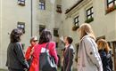 Eine Gruppe von Menschen steht in einem Innenhof und hört einem Führer zu. Alte Gebäude mit Fenstern und Pflanzen sind im Hintergrund zu sehen.