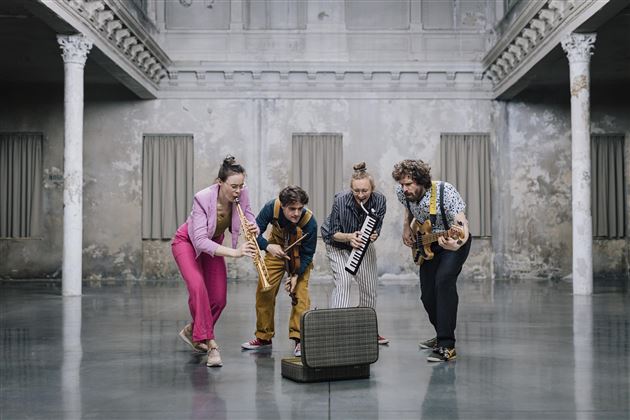 Een groep van vier muzikanten speelt in een grote, lege ruimte. Ze concentreren zich op een ouderwets radio en tonen veel plezier in de muziek.