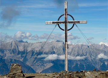 Ein großes Holzkreuz steht auf einem Berggipfel. Im Hintergrund sind majestätische Berge und ein blauer Himmel mit einigen Wolken sichtbar.