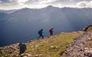 Deux randonneurs marchent sur une colline herbeuse avec un paysage montagneux impressionnant en arrière-plan. Le ciel est clair et le soleil brille à travers les nuages.
