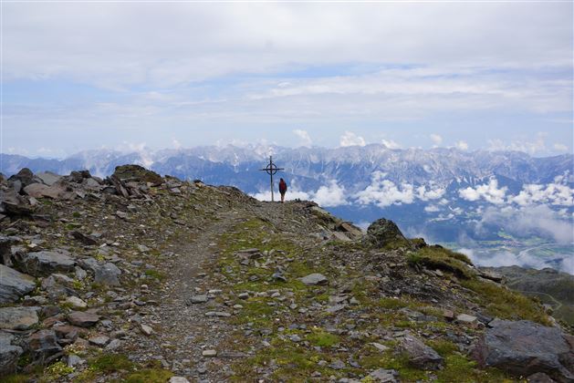 Un sentier de randonnée traverse un terrain rocheux avec vue sur les montagnes. En arrière-plan, des nuages se détachent dans un ciel dégagé.