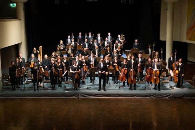Un orchestre complet se tient sur scène en tenue élégante. Les musiciennes et musiciens tiennent leurs instruments et regardent en direction du public.