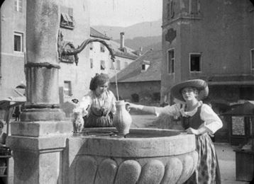 Two women in traditional clothing are standing by a fountain in a historic town. The surroundings are peaceful, with buildings in the background and a clear sky.