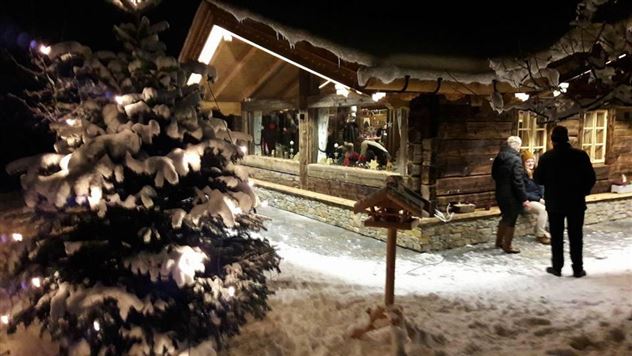 Un accogliente capanno di legno sotto un chiaro cielo notturno, circondato da alberi coperti di neve. Le luci brillano creando un'atmosfera festosa.