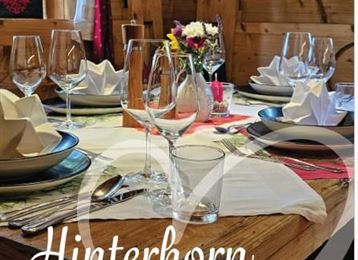 Eine elegant gedeckte Tafel in einer rustikalen Holzstube. Blumen und leuchtendes Geschirr verleihen dem Ambiente eine einladende Atmosphäre.