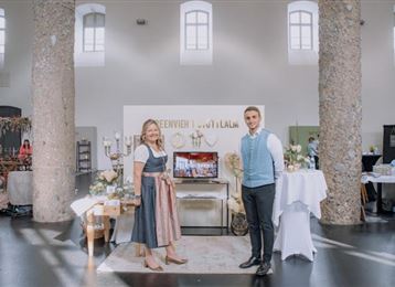 A festive room with two people smiling warmly at the camera. In the background, decorative elements and tables with flower arrangements can be seen.