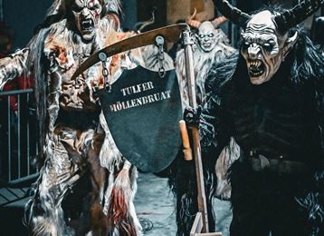 Un gruppo di figure spettrali con maschere a corna e costumi spaventosi. Portano uno scudo e sembrano partecipare a una festa o una parata.