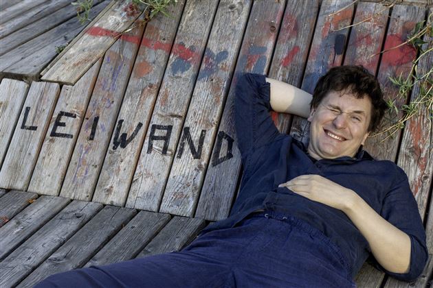 Un uomo sorridente tiene un microfono in mano. Sullo sfondo sono visibili punti di luce che formano un palco.