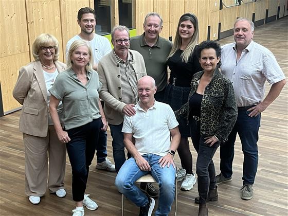 A group of ten people stands and sits smiling in a room. The atmosphere is friendly and the people are dressed in casual clothing.
