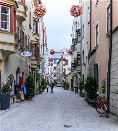 Eine schmale Gasse mit bunten Blumenbällen und historischen Gebäuden. Zwei Personen sind unterwegs, während Fahrräder und Pflanzen die Szene verschönern.