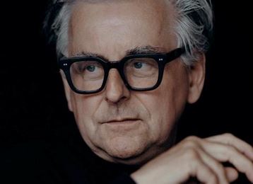 Un homme âgé avec des cheveux gris et des lunettes, qui regarde pensivement. Il porte un pull sombre et a une posture détendue.