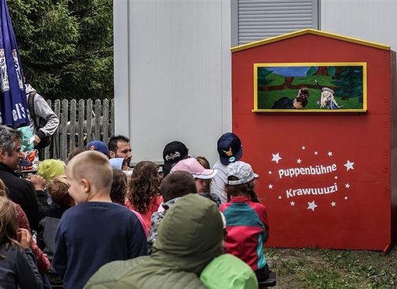 A puppet stage with a colorful backdrop attracts the children's attention. The audience sits eagerly in front of it and watches the puppet show.