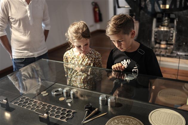 Zwei Kinder schauen fasziniert auf Objekte in einem Glasdisplay. Im Hintergrund ist eine erwachsene Person zu sehen.