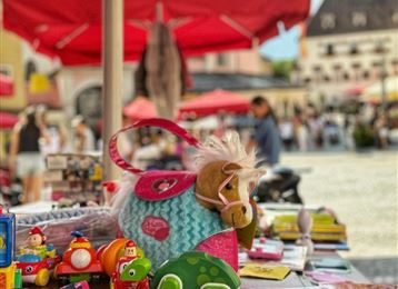 Ein Markttisch mit bunten Spielzeugen und einem Plüschpferd. Im Hintergrund sind Menschen und rote Sonnenschirme zu sehen.