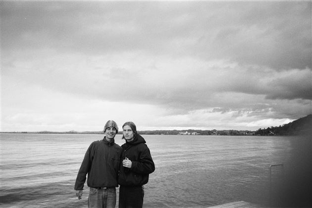 Two people are standing by the shore of a lake. The sky is cloudy and the water sparkles slightly.
