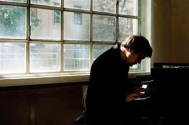 Ein Musiker spielt Klavier in einem hellen Raum. Sonnenlicht strömt durch ein großes Fenster.
