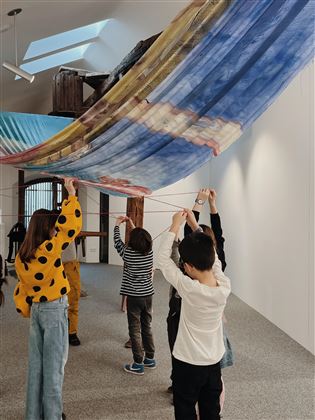 Eine Gruppe von Kindern zieht an bunten Tüchern in einem Ausstellungsraum. Die Atmosphäre ist kreativ und spielerisch.