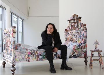 Une artiste est assise pensivement sur un lit coloré, décoré de différentes images. En arrière-plan, de grandes fenêtres et un environnement lumineux et minimaliste sont visibles.