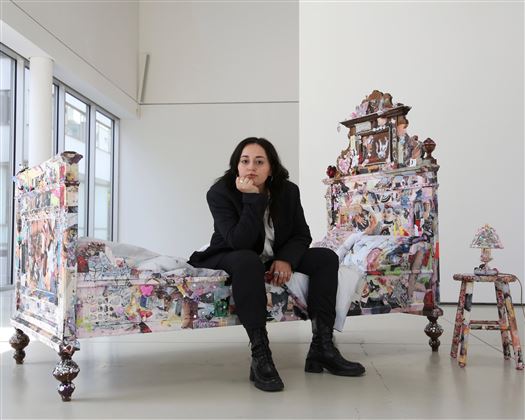 An artist is sitting pensively on a colorful bed adorned with various pictures. In the background, large windows and a bright, minimalist environment can be seen.