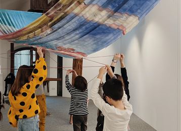 Een groep kinderen trekt aan kleurrijke doeken in een tentoonstellingsruimte. De sfeer is creatief en speels.
