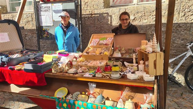 Un stand de marché avec des articles faits main et des décorations. Deux vendeurs se tiennent souriants derrière le stand.