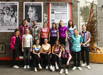 A group of young people stands in front of a building with posters. They are wearing colorful shirts and smiling at the camera.