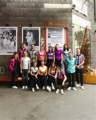 Een groep jongeren staat voor een gebouw met posters. Ze dragen kleurrijke shirts en glimlachen naar de camera.