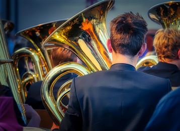 A group of musicians with several tubas in the foreground. The view from the back showcases the shiny instruments and part of the performance attire.