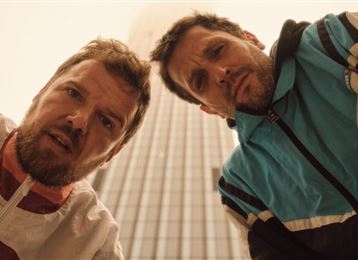 Deux hommes regardent l'objectif avec inquiétude. En arrière-plan, on peut voir un grand complexe de bâtiments.