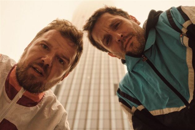 Twee mannen kijken bezorgd in de camera. Op de achtergrond is een hoog gebouwencomplex te zien.