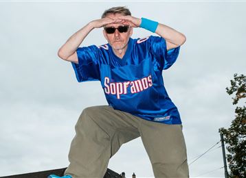 A man in a blue T-shirt with the inscription "Sopranos" poses with sunglasses. He has an athletic stance and is wearing baggy pants.