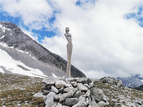 Een slanke houtsnede staat op een stenen ondergrond in een bergachtig landschap. Op de achtergrond zijn indrukwekkende bergen en een bewolkte lucht te zien.