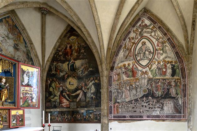 Eine prächtige Wand mit komplexen Fresken in einem historisch anmutenden Raum. Die Kunstwerke zeigen biblische Szenen und Figuren in lebendigen Farben.