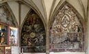 Eine prächtige Wand mit komplexen Fresken in einem historisch anmutenden Raum. Die Kunstwerke zeigen biblische Szenen und Figuren in lebendigen Farben.