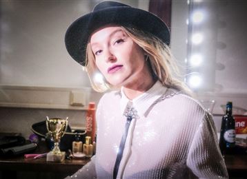 Une femme avec un chapeau pose dans une pièce élégante. En arrière-plan, des prix et des articles cosmétiques sont visibles.