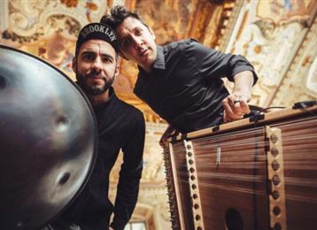 Two musicians are standing in a beautiful room with an elaborate ceiling. The man on the left is playing a handpan, while the other is presenting a different instrument.