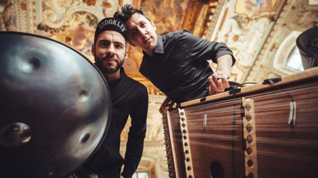 Deux musiciens se tiennent dans une belle pièce avec un plafond élaboré. L'homme à gauche joue d'un handpan, tandis que l'autre présente un autre instrument.