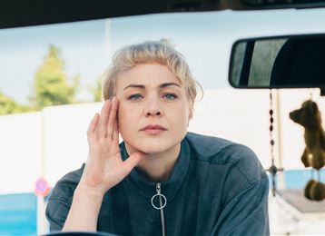 Een persoon zit aan de rand van een blauwe zwembad en kijkt in de camera. Ze draagt een casual shirt en heeft een glad kapsel.