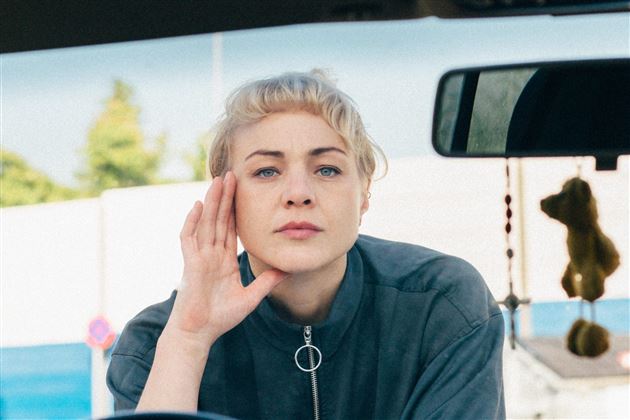 Une personne est assise au bord d'une piscine bleue et regarde la caméra. Elle porte une chemise décontractée et a une coiffure lisse.
